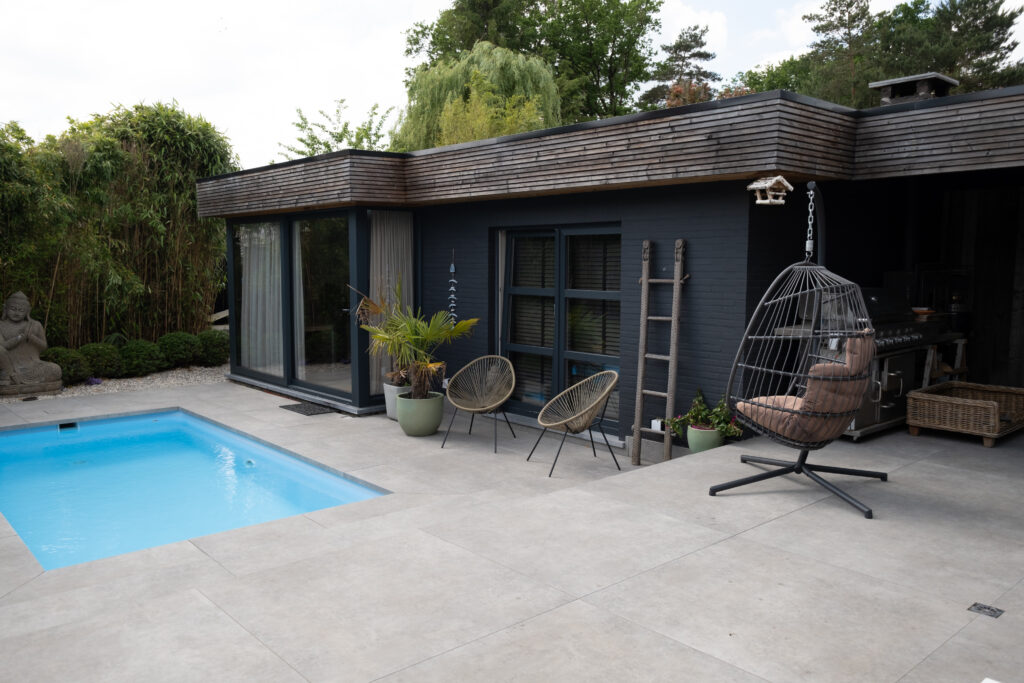 Terras en zwembadzone met London Mid keramische terrastegels in XXL-formaat 122×122 cm en bijpassende zwembadboorden in middengrijze tint. Antislip en kleurvaste tegels die naadloos aansluiten bij moderne gevel en ramen, voor een strak en elegant buitenproject.